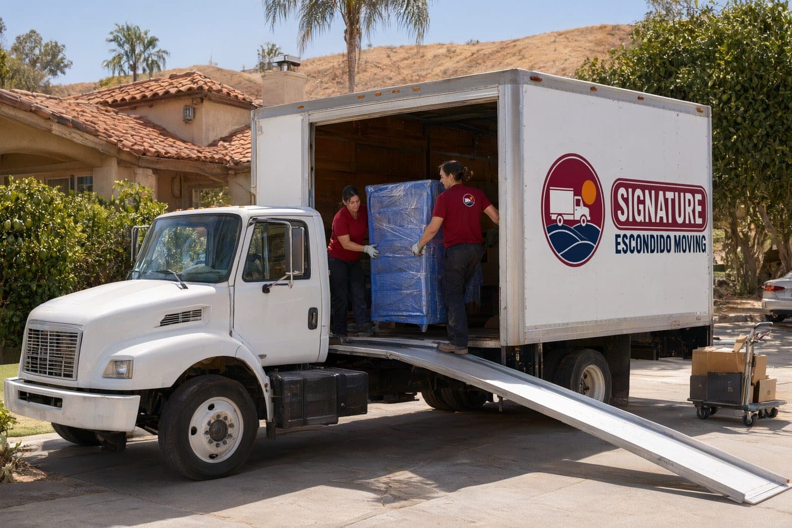 Two movers carrying a dresser wrapped in blue pads and stretch wrap up the ramp into our box truck